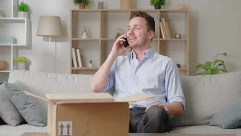 Man sit on sofa with parcel talks on smartphone thanks friend via phonecall for Stock Footage 169017708