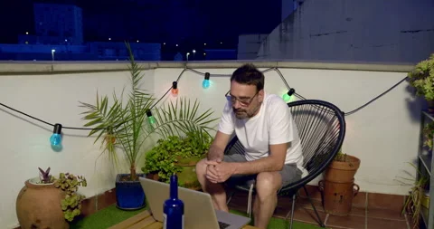 A man sits on a balcony at night, looking at a laptop. Stock Footage 336247146