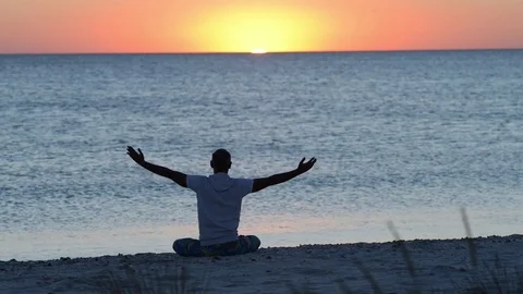Man sits on the beach next to surf, practicing yoga during amazing sunset Stock Footage 97152458