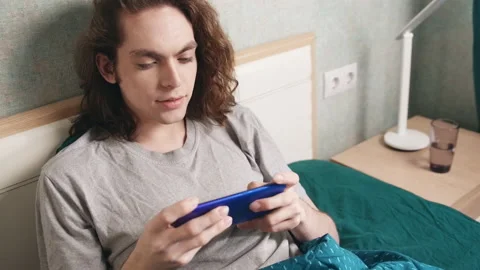 A man sits in bed and plays on a mobile phone, he smiles then gets upset Stock-Footage 196812699