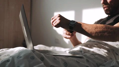 A man sits on a bed covered with a blanket puts on a wristwatch and works on a Stock Footage 154342444