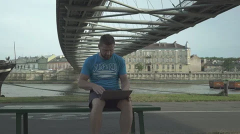 Man sits on the bench and starts typing on the laptop, pan shot. Vidéo 65205209