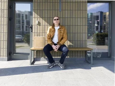 Man sits on bench with cardboard box after purchase near building. Person res Stock Photos