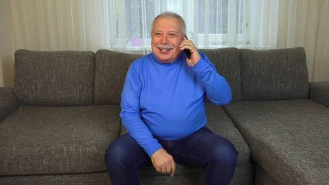 Man sits on brown sofa holds black cellphone and talks Stock Footage 108605044