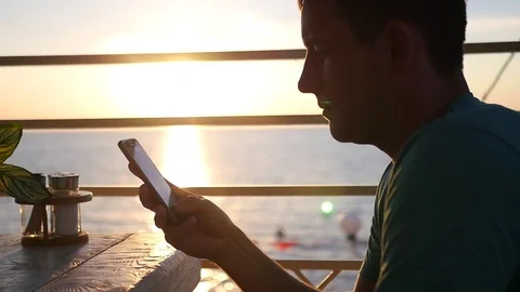 A man sits in a cafe on the background of a beautiful sunset, with a phone in Stock Footage 79928441