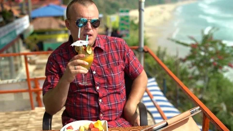 A man sits in a cafe at a table by the sea drinking a cocktail Stock Footage 74761688