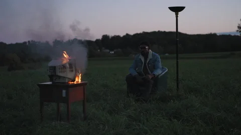 Man sits calmly while smoke and fire rise from a burning appliance in a field Stock Footage 319969905