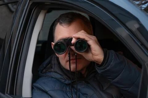 A man sits in a car looking through binoculars out of the vehicle window, c.. Фото