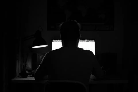 A man sits at a computer in a room at a table at night with blue lighting and Stock Photos