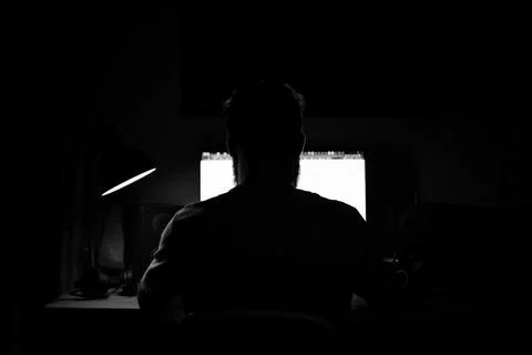 A man sits at a computer in a room at a table at nigh programming black and w Stock-Fotos