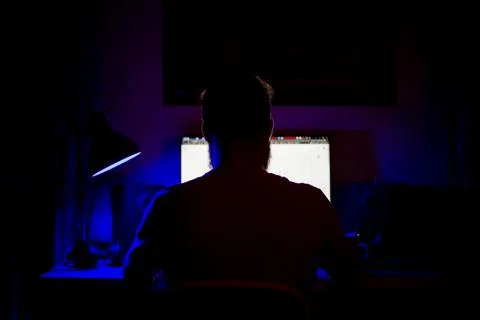 A man sits at a computer in a room at a table at night with blue lighting and Photos