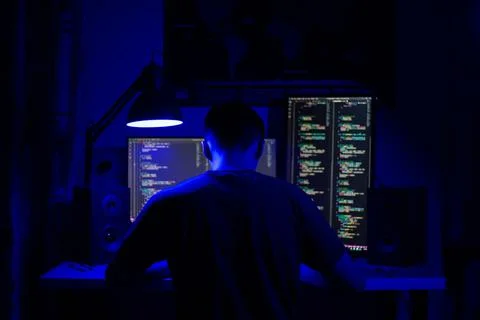 A man sits at a computer in a room at a table at night with blue lighting and Stock Photos