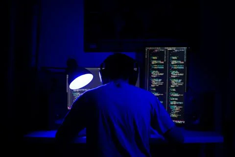 A man sits at a computer in a room at a table at night with blue lighting and Foto stock