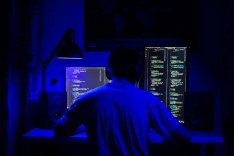 A man sits at a computer in a room at a table at night with blue lighting and Fotos de archivo
