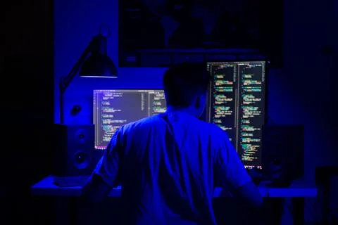 A man sits at a computer in a room at a table at night with blue lighting and Stock-Fotos