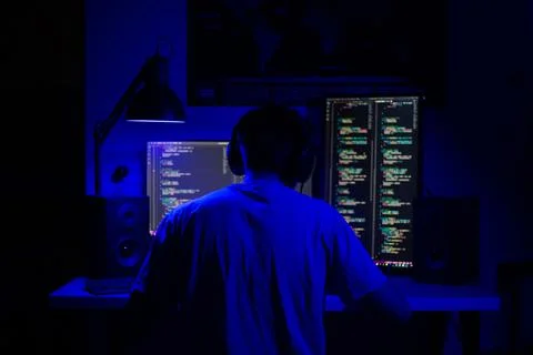 A man sits at a computer in a room at a table at night with blue lighting and Stock-Fotos