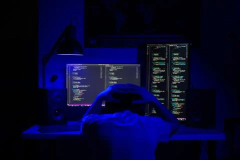 A man sits at a computer in a room at a table at night with blue lighting and Stock-Fotos