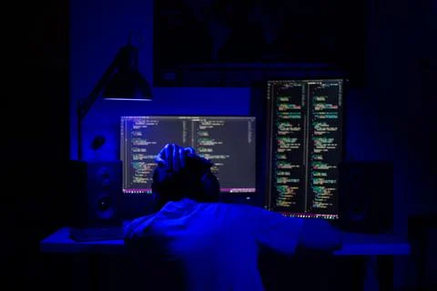 A man sits at a computer in a room at a table at night with blue lighting and Stock-Fotos