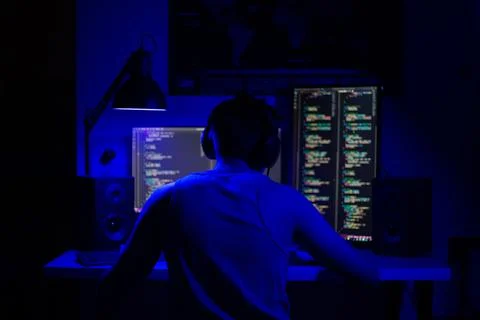 A man sits at a computer in a room at a table at night with blue lighting and Stock Photos