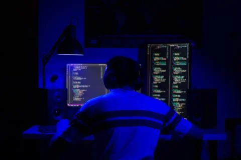 A man sits at a computer in a room at a table at night with blue lighting and Stock-Fotos