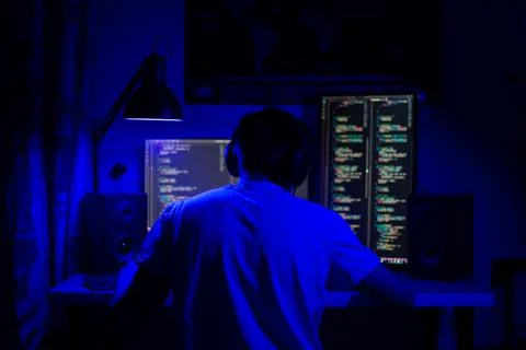 A man sits at a computer in a room at a table at night with blue lighting and Stock-Fotos