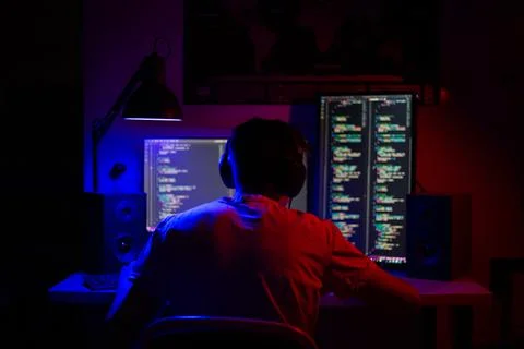 A man sits at a computer in a room at a table at night with blue lighting and Stock Photos