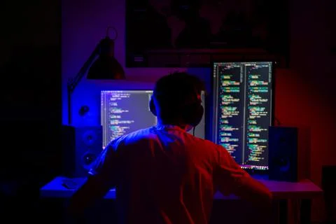 A man sits at a computer in a room at a table at night with blue lighting and Stock-Fotos