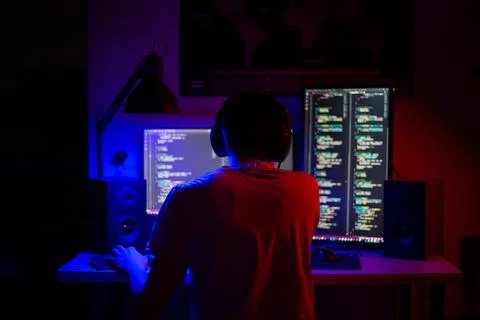 A man sits at a computer in a room at a table at night with blue lighting and Stock-Fotos