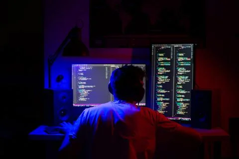 A man sits at a computer in a room at a table at night with blue lighting and Stock-Fotos