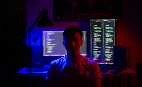 A man sits at a computer in a room at a table at night with blue lighting and Stock Photos
