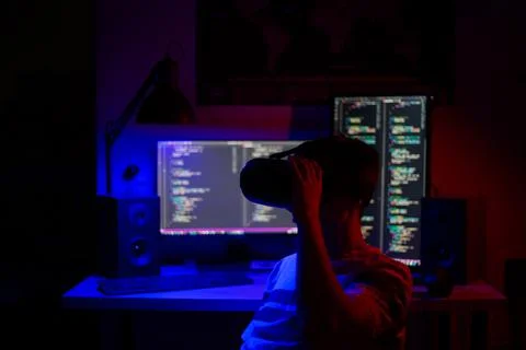 A man sits at a computer in a room at a table at night with blue lighting and Stock-Fotos