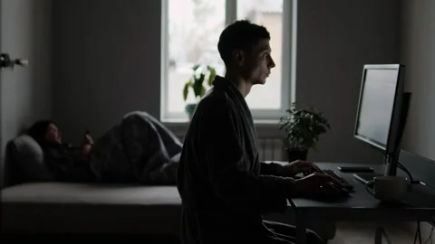 Man sits at computer while woman scrolls phone in bed, distant and disconnected Stock Footage 306390053