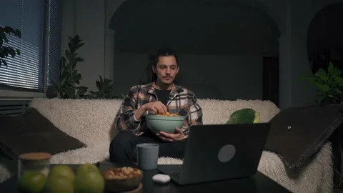 A man sits on the couch, chooses a video to watch, picks up a cup of chips Vidéo 170690488