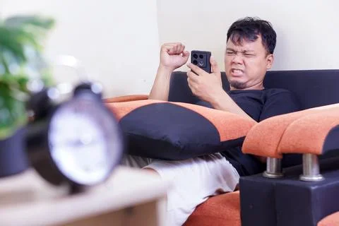A man sits on a couch, frustrated while looking at his smartphone, with an .. Stock Photos