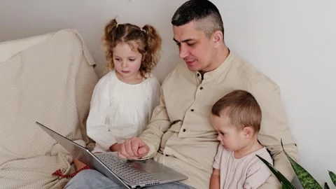 A man sits on a couch with two children. He helps them use a laptop while they Vídeo Stock 336459852