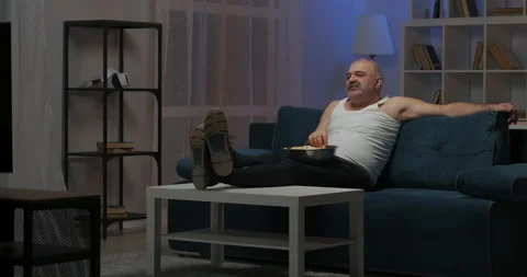 A man sits on the couch watching TV, threw his legs on the table and eats Stock Footage 148197114