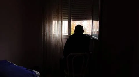 A man sits in a dark room and looks out of an open window Stock Photos