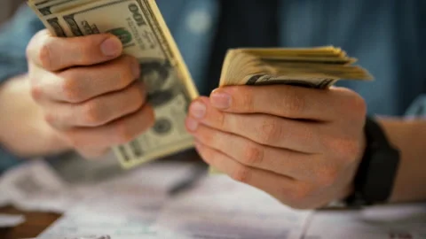 A man sits at a desk meticulously counting cash while referring to detailed Stock Footage 338186113