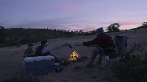 Man Sits Down By Campfire, His Friend Grabs A Beer From A Cooler For Him Stock Footage