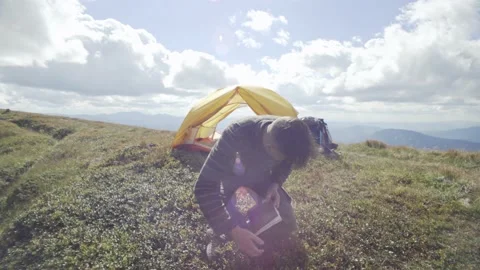 A man sits down to read a book near a tent while hiking in the mountains Stock Footage 217397085