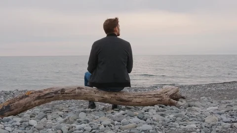 A man sits on a driftwood on the beach  throws stones into the sea. Video stock 127082754