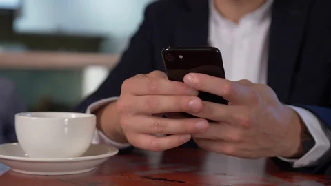 A man sits drinking coffee / tea at a table and performs actions on his phone. Stock Footage 106001236
