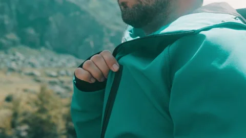 A man sits on the edge of a cliff in a green anorak and takes out his glasses Stock Footage 166065642
