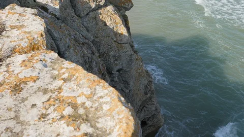 Man sits on the edge of a cliff. Waves wash the sandy shore. Ridge is broken on Stock-Footage 99704315