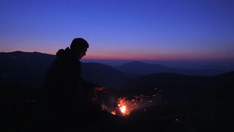 A man sits in the evening by a campfire in the mountains and warms his hands  Stock Footage 135816030