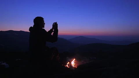 A man sits in the evening by a fire in the mountains and takes pictures of th Stock Footage 135815992