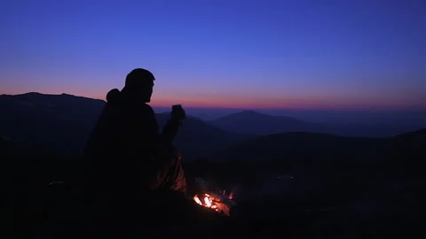 A man sits in the evening by a fire in the mountains and drinks a hot drink f Stock Footage 135816073
