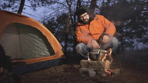 A man sits by the fire with a kettle in which water boils. Video stock 104409722