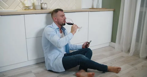 A man sits on the floor in the kitchen, drinks red wine and reads messages on Stock Footage 146884722
