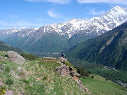 A man sits in front of a cliff on the edge of a mountain and looks at the huge Stock Photos